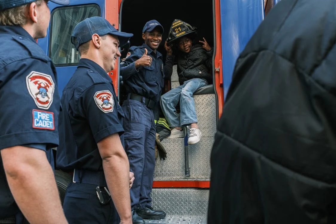 Fire cadets in uniform stand beside an engine while a child tries on a firefighter's helmet in the doorway