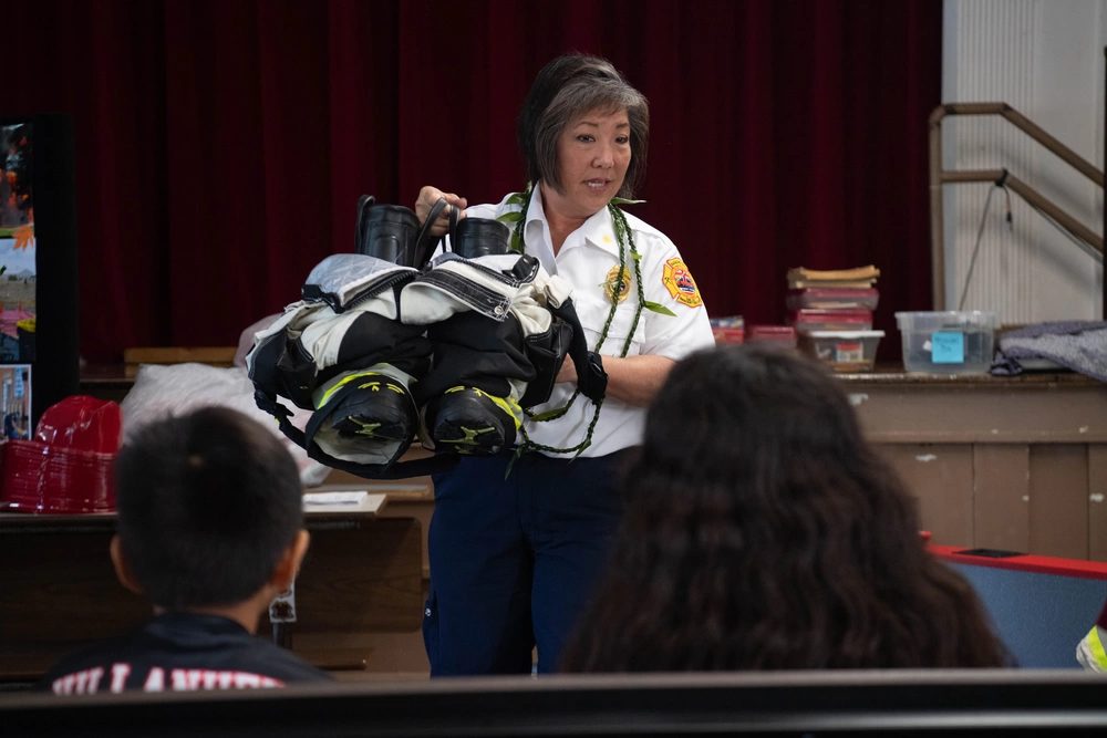 A fire chief holds up a set of turnout gear while speaking to students at a school assembly