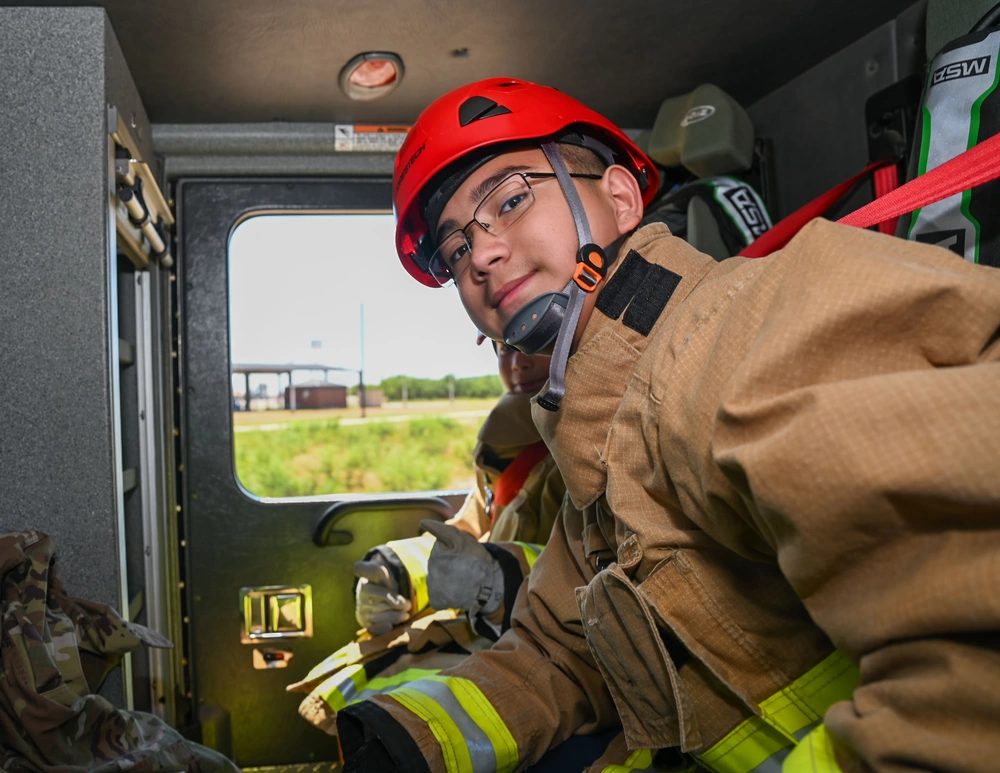 A young firefighter in turnout gear and a red helmet seated in the crew cab of a fire engine